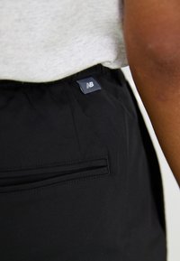 Pantalons de sport noirs avec une texture lisse, une taille élastique et une petite étiquette logo près de la poche arrière. Design simple et fonctionnel.
