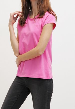 Mujer con camiseta rosa brillante de manga corta y vaqueros negros, con una mano tocándose el cabello y la otra apoyada en la cintura.