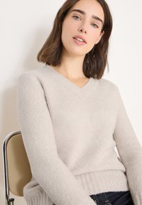Hellgrauer, strukturierter V-Ausschnitt-Pullover mit geripptem Saum und langen Ärmeln. Weiches Strickmaterial; taillierte Silhouette; über dunkler Hose getragen.