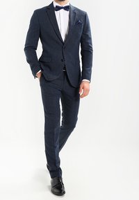 Granatowy garnitur z mieszanki wełny o dopasowanym kroju, jednorzędowa marynarka z klapami wciętymi oraz spodnie dopasowane. Do kompletu biała koszula i muszka.