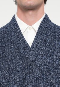 Jūras zilas krāsas adīts džemperis ar V-veida izgriezumu, ribota tekstūra un vidēja biezuma, valkāts virs baltas apkaklītes krekla.