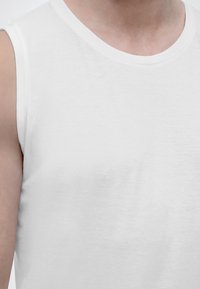 Κοντινό πλάνο ενός ατόμου που φοράει απλή λευκή αμάνικη μπλούζα, εμφανίζοντας το άνω μέρος του στήθους, τον λαιμό και τον αριστερό ώμο.