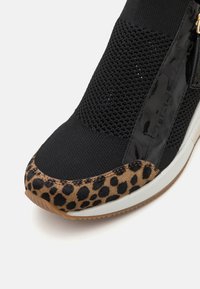 Baskets noires avec une tige texturée, des accents brillants, et un bout en imprimé léopard, dotées d'une semelle blanche et de coutures complexes.