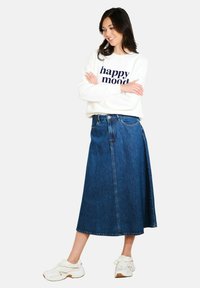 Donkerblauwe denim A-lijn rok met een voorzicht, gecombineerd met een witte sweatshirt met marineblauwe tekst. Model draagt witte sneakers.