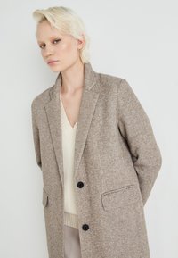 Hnedý texturovaný vlnový kabát s elegantným strihom, s klopami a čiernymi gombíkmi, preložený cez krémovo sfarbený pletený top.