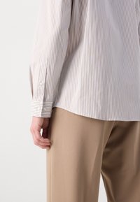 White and tan striped button-up shirt with long sleeves, featuring a cuffs with buttons. Paired with beige, relaxed-fit trousers.