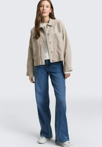 Vrouw met lang bruin haar, een beige jasje met knopen, een wit shirt, blauwe wijde jeans en witte sneakers, staand tegen een effen achtergrond.