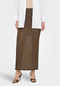 Bruine doorschijnende maxi-rok met een geribde textuur en decoratief patroon, gecombineerd met een beige top en een witte button-up blouse.