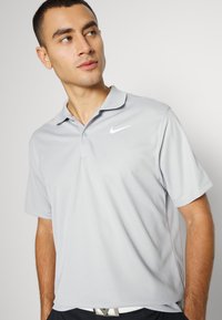Sivá Nike polo majica s klasičnim ovratnikom, kratkimi rokavi in belim logotipom na levi strani prsnega dela. Gladka, lahka tkanina.