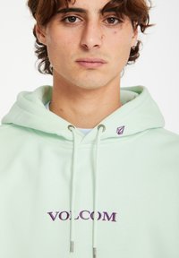 Mintgroene hoodie met trekkoord, voorzien van een paarse geborduurde "VOLCOM"-logo en een klein logoaccent bij de halslijn.