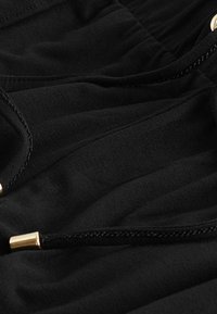 Détail en tissu noir avec une texture douce, un design élastique et des accents en métal doré. Couture visible le long des coutures et des bords.