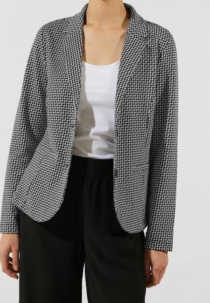 Person, die einen schwarz-weißen Hahnentritt-Blazer über einem weißen Tanktop und schwarzen Hosen trägt, vor einem einfarbigen Hintergrund steht.