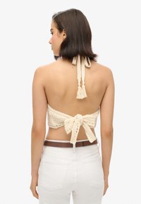 Gehaakte haltertop in crème, met een gestrikte achterkant en franjerandjes. Gekozen in combinatie met witte high-waisted broek.