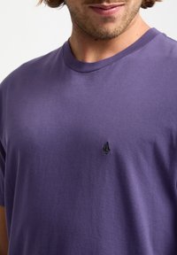 Lila kurzärmliges T-Shirt aus weicher Baumwolle mit Rundhalsausschnitt. Verfügt über ein kleines schwarzes gesticktes Logo auf der Brust. Glatte Textur.