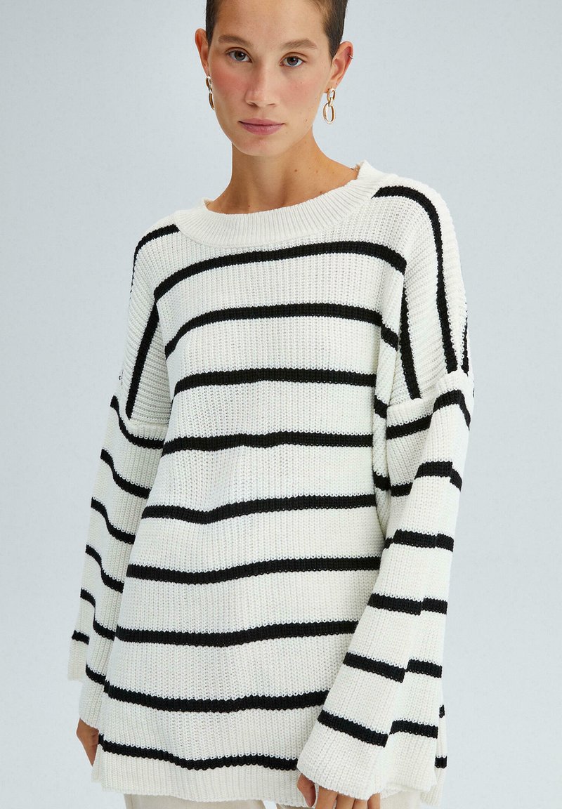 Pull en maille blanc avec des rayures noires horizontales, coupe ample, manches larges et encolure côtelée. Texture douce avec un design décontracté.
