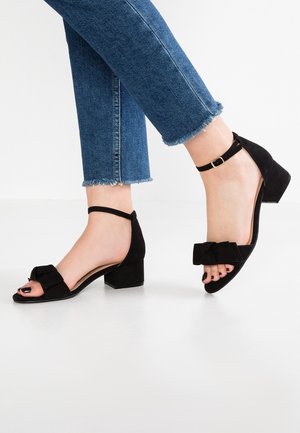 Fødder iført sorte block-hælede sandaler med ankelremme og sløjfer, kombineret med blå jeans med frynset kant på en hvid gulv.