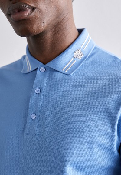 Polo de cor azul claro feito de tecido texturizado, com uma pala de dois botões, emblema bordado no colarinho e detalhes em riscas brancas.