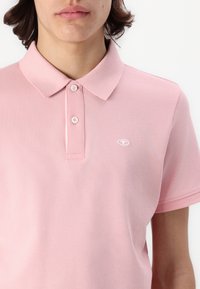 Lichtroze polohemd met een geweven structuur, voorzien van een kraag en een knoopsluiting met twee knopen. Klein geborduurd logo op de linkerborst.