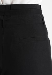 Zwarte broek met een gladde textuur, brede tailleband en een enkele achterzak. Kenmerkt zich door nette stiksels en minimalistische designelementen.
