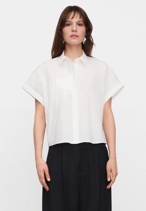 Kobieta ubrana w białą koszulę na guziki z krótkim rękawem i delikatnym haftem oraz czarne, szerokie spodnie z wysokim stanem, stojąca na białym tle.