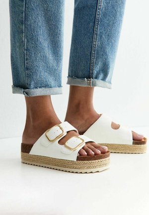 Stopy w białych sandałach na platformie z złotymi klamrami i plecionymi podeszwami, zestawione z podwiniętymi niebieskimi jeansami.