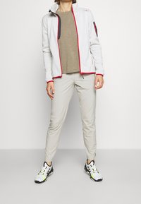Giacca in pile grigia chiara con dettagli rossi e zip nera, abbinata a pantaloni beige e sneakers bianche con dettagli neri e verde neon.