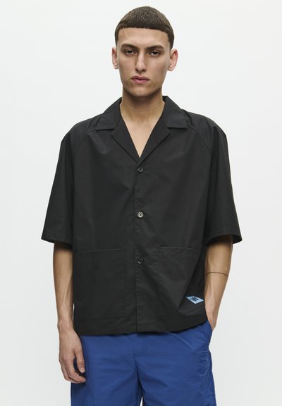 Black short-sleeve shirt with a notch collar, two front pockets, and a blue logo accent. Button-down closure and smooth fabric texture.