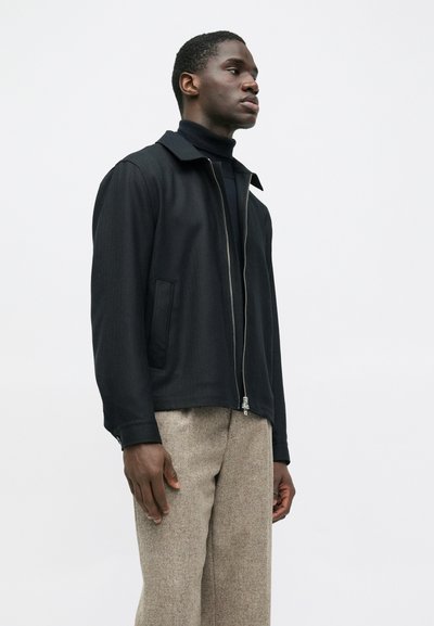 Vīrietis, tērpies melnā jakā ar rāvējslēdzēju virs melna turtlenecka un smilškrāsas teksturētām biksēm, stāv uz nekrāsotas baltas fona.