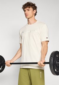 Beige Baumwoll-T-Shirt mit einem reflektierenden "Champion"-Logo auf der Brust. Kurze Ärmel, lässige Passform, kombiniert mit grünen Jogginghosen.