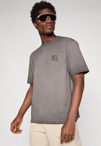 Szary bawełnowy t-shirt o luźnym kroju i krótkich rękawach. Ma małe czarne logo na piersi. Noszony z dużymi okularami przeciwsłonecznymi.