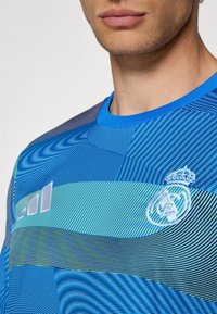 Blauwe sportieve shirt met mesh textuur, voorzien van lijnen in verschillende tinten, en een geborduurd logo op de borst.