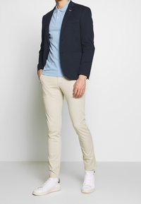 Marineblazer over een lichtblauw polo, gecombineerd met beige slim-fit broek en witte sneakers. Kenmerkt zich door een strak ontwerp en casual pasvorm.