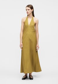 Γυναίκα όρθια, φοράει μίντι φόρεμα χακί σε στυλ halter-neck χωρίς μανίκια και μαύρα επίπεδα παπούτσια, σε απλό ανοιχτόφωτο φόντο.