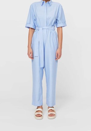 Combinaison courte à manches courtes bleu clair, boutonnée, avec une taille nouée, des jambes droites et des poches latérales. Portée avec des sandales beige.