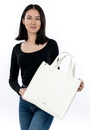 Junge Frau mit schulterlangem dunklem Haar, trägt ein schwarzes Oberteil und Jeans, hält eine große weiße Tragetasche mit zwei Griffen.