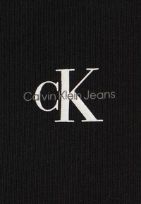 Černá tkanina Calvin Klein Jeans s bílým logem. Logo jasně zobrazuje "cK" s nápisem "Calvin Klein Jeans" pod ním.