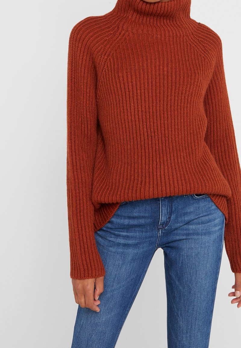 Rust orange knitted sweater with a high collar, ribbed texture, and side slits, paired with blue jeans.