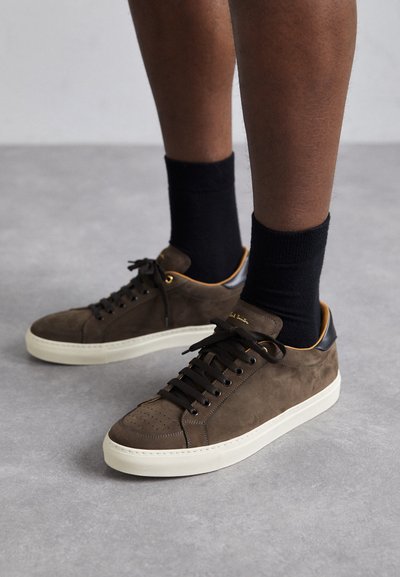 Sneaker in suede marrone con dettagli in pelle nera, suole color crema e lacci marroni. Presentano una punta perforata e un dettaglio in logo dorato sul lato.