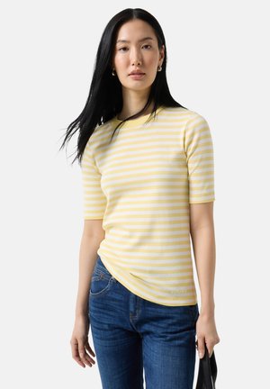 Ung kvinde med langt sort hår iført en gul og hvidstribet kortærmet top og blå jeans, der holder en sort taske.