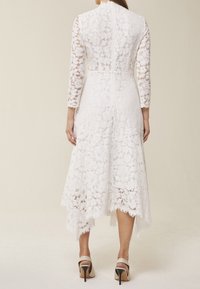 Abito bianco in pizzo con maniche lunghe, scollatura alta e orlo svasato. Presenta motivi floreali e una finitura testurizzata. I tacchi completano l'outfit.