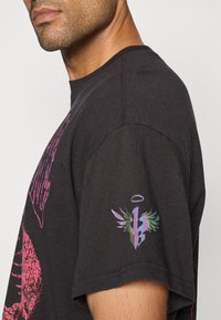 Sort bomulds t-shirt med lyserøde grafiske tryk og et flerfarvet logo med vinger på højre ærme. Korte ærmer, rund halsudskæring.
