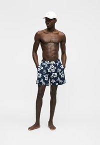 Hombre sin camiseta vistiendo pantalones cortos de baño azul marino con patrones florales blancos y de leopardo, y una gorra blanca de béisbol, de pie con las manos en los bolsillos.