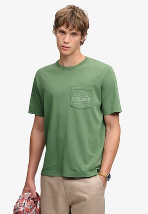 Jeune homme portant un t-shirt vert Superdry et un pantalon beige, tenant une chemise à carreaux rouge dans une main, avec une montre au poignet.
