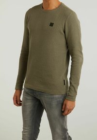 Oliwkowy sweter z długim rękawem i teksturowanym wzorem, okrągły dekolt oraz mała łatka z logo na piersi, zestawiony z szarymi dżinsami.