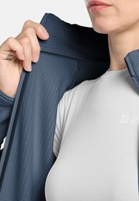 Veste texturée bleu marine avec fermeture éclair à l'avant, portée sur une chemise ajustée gris clair avec un logo. Accent sur le design du col et des manches.