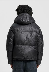 Person med mørkt krøllet hår iført en sort puffjakke med hætte, vendt væk, stående mod en ensfarvet lys baggrund.