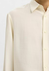 Camicia color crema realizzata in un tessuto liscio, con chiusura a bottoni, colletto a punta e texture sottile. Presenta piccoli bottoni e maniche lunghe.