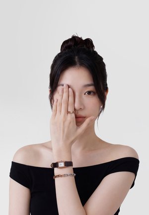 Fiatal nő sötét hajjal, kontyba fogva, fekete vállatlan felsőt visel, kezével eltakarja az egyik szemét, miközben órát és karkötőt mutat.