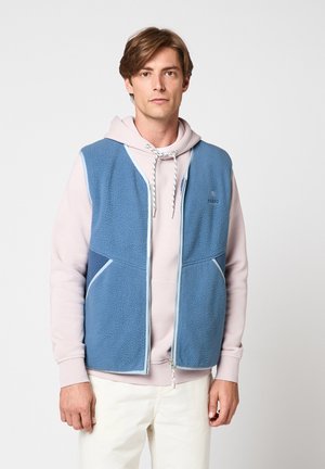 Jeune homme aux cheveux châtain clair portant un sweat à capuche rose clair et un gilet en polaire bleu zippé, debout devant un fond clair uni.