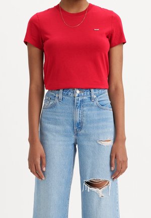 Personne portant un t-shirt rouge à manches courtes avec un petit logo et un jean taille haute bleu clair avec des trous déchirés aux deux jambes.